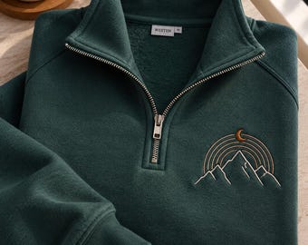 May include: A dark green quarter-zip sweatshirt featuring a silver metal zip and a ribbed collar. An embroidered design on the chest depicts a mountain range with concentric semicircular lines above it and a small crescent moon at the top.