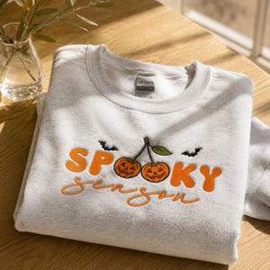 May include: A light gray crewneck sweatshirt featuring embroidered orange text that reads "SPOOKY season" with two orange jack-o'-lanterns on stems resembling cherries in the center, flanked by two small black embroidered bats.