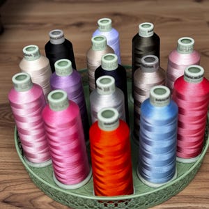 May include: A collection of fifteen cylindrical spools of Madeira embroidery thread in various colors, including red, orange, blue, pink, purple, black, and beige, arranged upright on a round, light green metal tray with a decorative lattice edge.