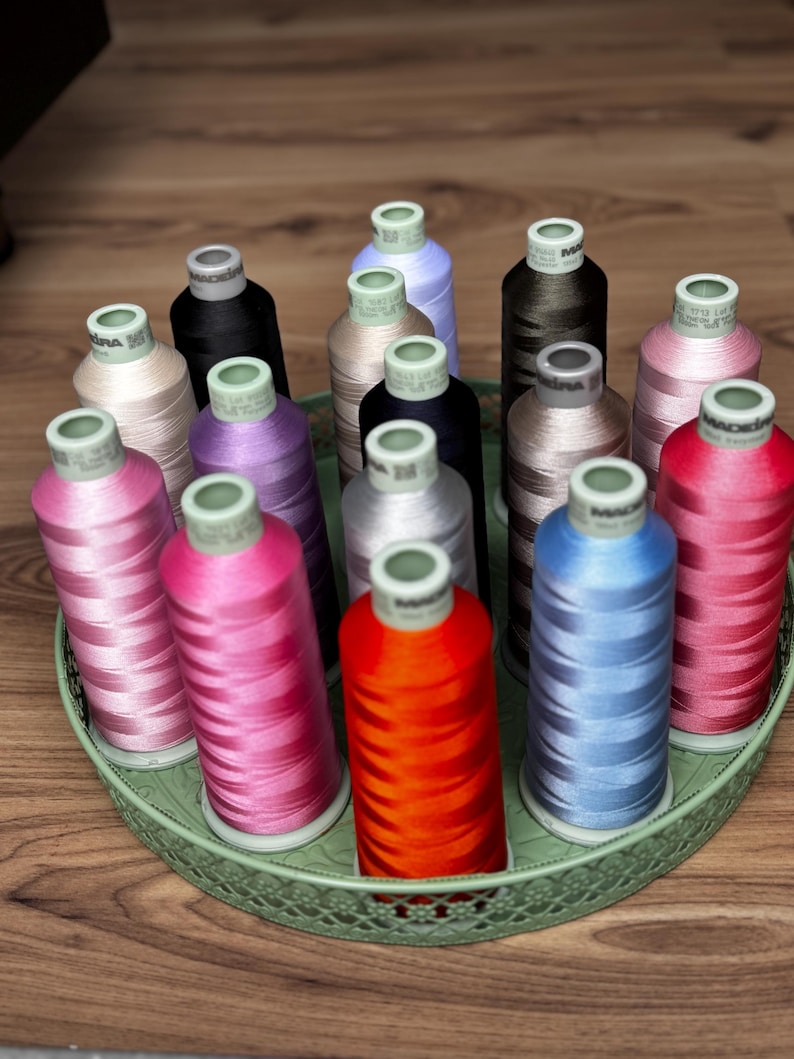 Pode incluir: Uma cole&ccedil;&atilde;o de quinze carret&eacute;is cil&iacute;ndricos de linha de bordar Madeira em v&aacute;rias cores, incluindo vermelho, laranja, azul, rosa, roxo, preto e bege, dispostos verticalmente numa bandeja redonda de metal verde-claro com borda decorativa.