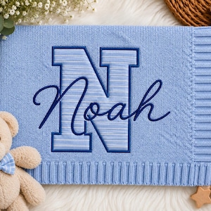 Könnte beinhalten: Eine hellblaue Strickdecke mit einem großen, dunkelblau bestickten Buchstaben N, der ein gestreiftes Muster aufweist. Der Name Noah ist in dunkelblauer Schreibschrift über den Buchstaben gestickt. Ein beiger Teddybär mit blauer Fliege ist zu sehen.