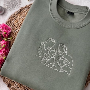 May include: An olive green crewneck sweatshirt featuring a white line art embroidery of a family of four on the center chest. The garment has a ribbed collar and long sleeves, showcasing a minimalist outline design of two adults and two children.