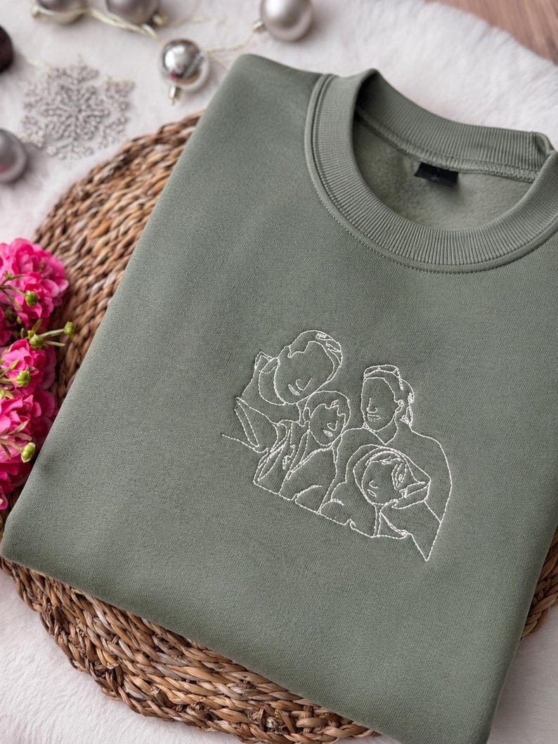 May include: An olive green crewneck sweatshirt featuring a white line art embroidery of a family of four on the center chest. The garment has a ribbed collar and long sleeves, showcasing a minimalist outline design of two adults and two children.