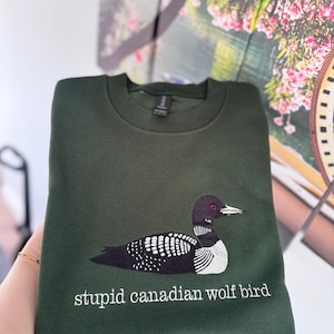 Pode incluir: Uma camisola de gola redonda em verde floresta escuro com um bordado de um mergulh&atilde;o com detalhes a preto, branco e vermelho. Por baixo da ave, o texto "stupid canadian wolf bird" est&aacute; bordado em letras min&uacute;sculas brancas com serifa.