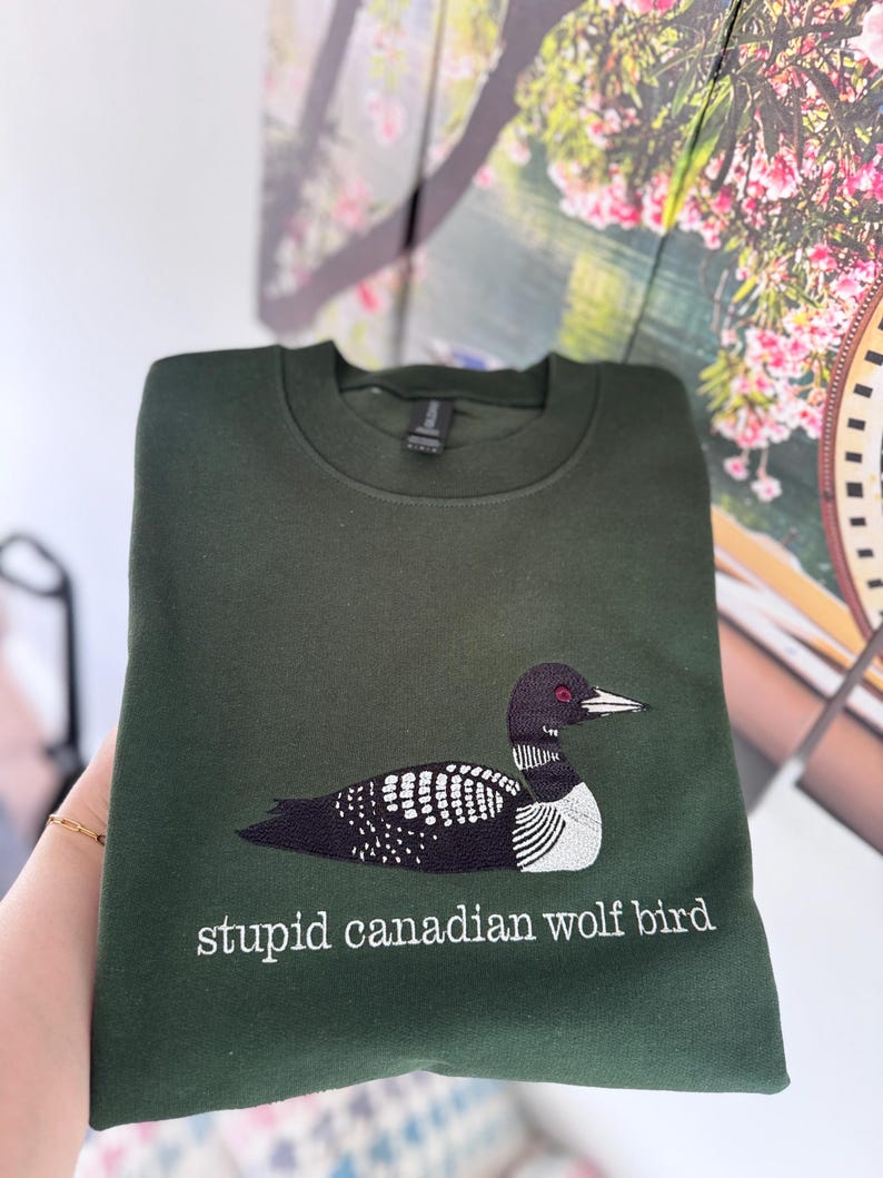 Pode incluir: Uma camisola de gola redonda em verde floresta escuro com um bordado de um mergulh&atilde;o com detalhes a preto, branco e vermelho. Por baixo da ave, o texto "stupid canadian wolf bird" est&aacute; bordado em letras min&uacute;sculas brancas com serifa.