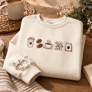 Pode incluir: Uma camisola de gola redonda cor de creme com bordados castanhos de ícones relacionados com café, incluindo um copo, grãos, uma chávena, uma máquina e um saco. A manga tem bordado o nome Sofia e um pequeno coração em fio castanho.