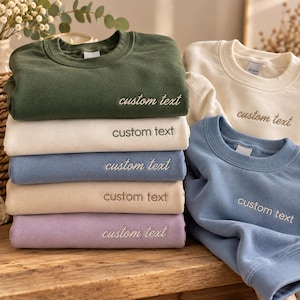 Peut inclure: Une pile de cinq sweat-shirts à col rond en vert olive, crème, bleu poussiéreux, beige et lavande, avec deux autres sweat-shirts en crème et bleu. Chaque vêtement présente le texte brodé custom text sur la poitrine.