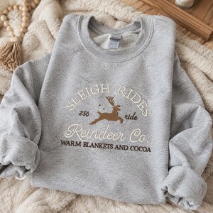 May include: A light gray crewneck sweatshirt featuring embroidered text that reads SLEIGH RIDES, Reindeer Co., 25&cent;, ride, and WARM BLANKETS AND COCOA. A brown embroidered reindeer is centered between the text, accompanied by small star accents.
