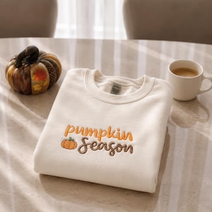 Puede incluir: Una sudadera de cuello redondo color crema con texto bordado que dice "pumpkin season" en hilo naranja y marrón, acompañada de un pequeño icono de calabaza naranja bordado situado a la izquierda de la palabra season.