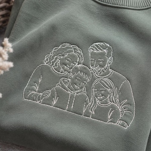 May include: An olive green crewneck sweatshirt featuring a white line-art embroidery of a family of four. The design depicts two adults and two children positioned closely together, showcasing detailed facial features and clothing outlines.