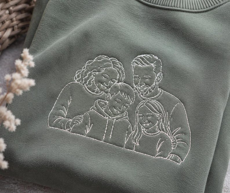 May include: An olive green crewneck sweatshirt featuring a white line-art embroidery of a family of four. The design depicts two adults and two children positioned closely together, showcasing detailed facial features and clothing outlines.