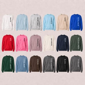 May include: A collection of eighteen crewneck sweatshirts displayed in a grid, each featuring a unique solid color including white, ash grey, sport grey, sky blue, sand, royal blue, red, pink, paragon, off white, navy, military green, forest green, stone blue, cocoa, dark heather, black, and charcoal, with the color name printed vertically in black text on the right side of each garment.
