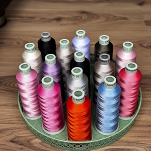 May include: A collection of fifteen Madeira embroidery thread spools in various colors including red, orange, pink, blue, purple, brown, black, and beige, arranged upright on a round, light green, decorative metal tray with a lattice edge.