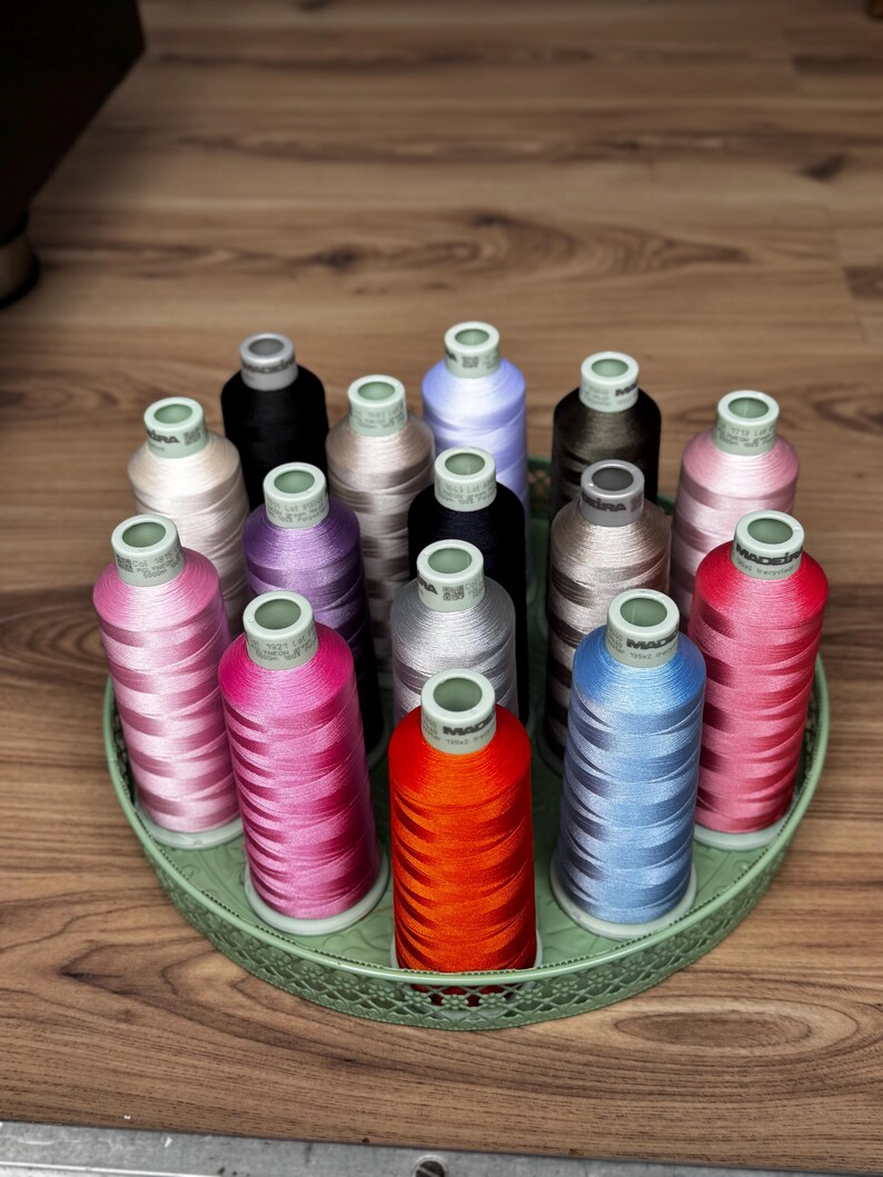 May include: A collection of fifteen Madeira embroidery thread spools in various colors including red, orange, pink, blue, purple, brown, black, and beige, arranged upright on a round, light green, decorative metal tray with a lattice edge.