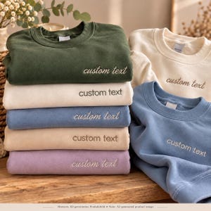 May include: A stack of five crewneck sweatshirts in olive green, cream, dusty blue, beige, and lavender, plus two additional sweatshirts in cream and blue. Each garment features the embroidered text custom text on the chest.