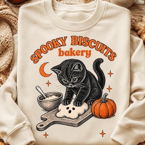 Pode incluir: Uma camisola de gola redonda cor de creme com um gráfico de um gatinho preto a amassar massa numa tábua de madeira. O design inclui uma lua crescente laranja, uma abóbora, uma taça e o texto SPOOKY BISCUITS bakery.