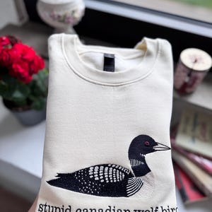 Pode incluir: Uma camisola de gola redonda cor de creme com um bordado de um mergulh&atilde;o em preto e branco. Por baixo da ave, o texto "stupid canadian wolf bird" est&aacute; bordado em letras min&uacute;sculas pretas. A pe&ccedil;a tem gola e punhos canelados.
