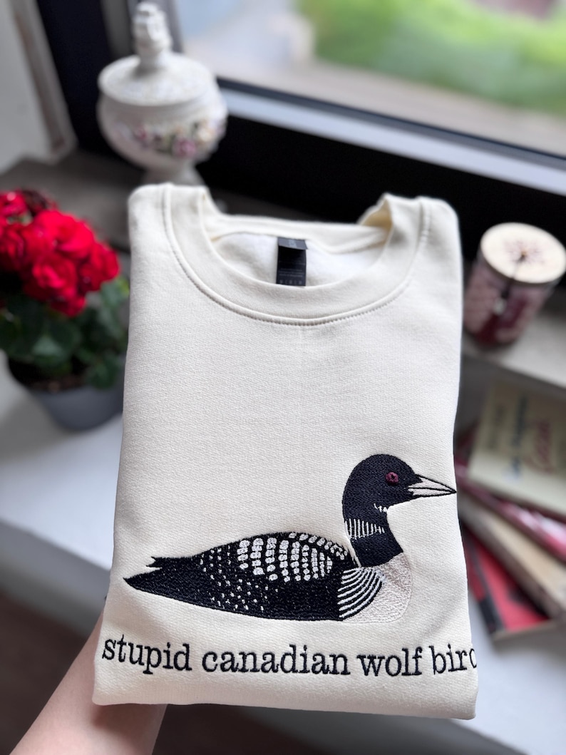 Pode incluir: Uma camisola de gola redonda cor de creme com um bordado de um mergulh&atilde;o em preto e branco. Por baixo da ave, o texto "stupid canadian wolf bird" est&aacute; bordado em letras min&uacute;sculas pretas. A pe&ccedil;a tem gola e punhos canelados.