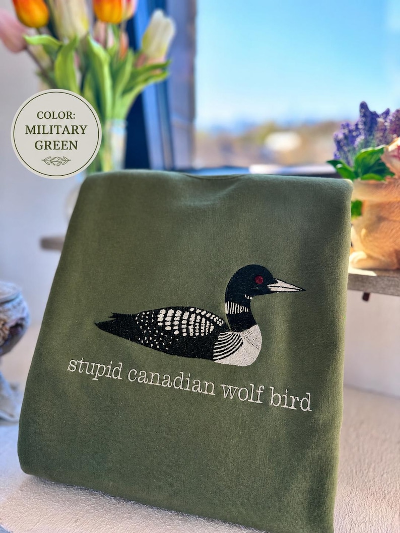 Pode incluir: Uma camisola verde militar com um bordado de um mergulh&atilde;o preto e branco com um olho vermelho. Por baixo da ave, o texto "stupid canadian wolf bird" est&aacute; bordado em fio branco. Uma etiqueta circular indica "COLOR: MILITARY GREEN".