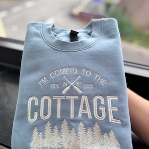 Könnte beinhalten: Ein hellblaues Sweatshirt mit Rundhalsausschnitt und weißer Stickerei. Der Text lautet I'M COMING TO THE COTTAGE, ESTD 24 81 2025 und CANADA. Das Design zeigt gekreuzte Ruder und einen Nadelwald, die in weißem Garn auf die Vorderseite gestickt sind.
