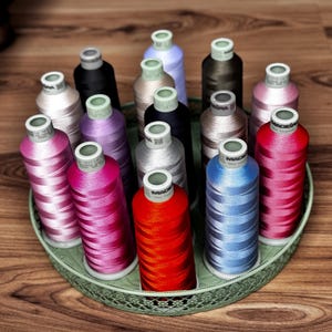 May include: A collection of fifteen cylindrical spools of Madeira embroidery thread in various colors, including red, blue, pink, purple, black, and grey, arranged upright in a round, light green decorative metal tray with a lattice pattern.