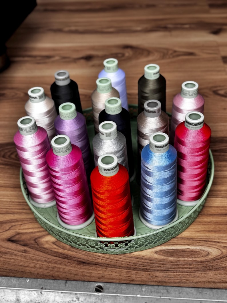 May include: A collection of fifteen cylindrical spools of Madeira embroidery thread in various colors, including red, blue, pink, purple, black, and grey, arranged upright in a round, light green decorative metal tray with a lattice pattern.
