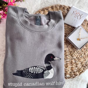 Pode incluir: Uma camisola cinzenta de gola redonda com um bordado de um mergulh&atilde;o em preto e branco. Por baixo da ave, o texto "stupid canadian wolf bird" est&aacute; bordado a branco. A camisola tem um interior em polar e uma gola padr&atilde;o.
