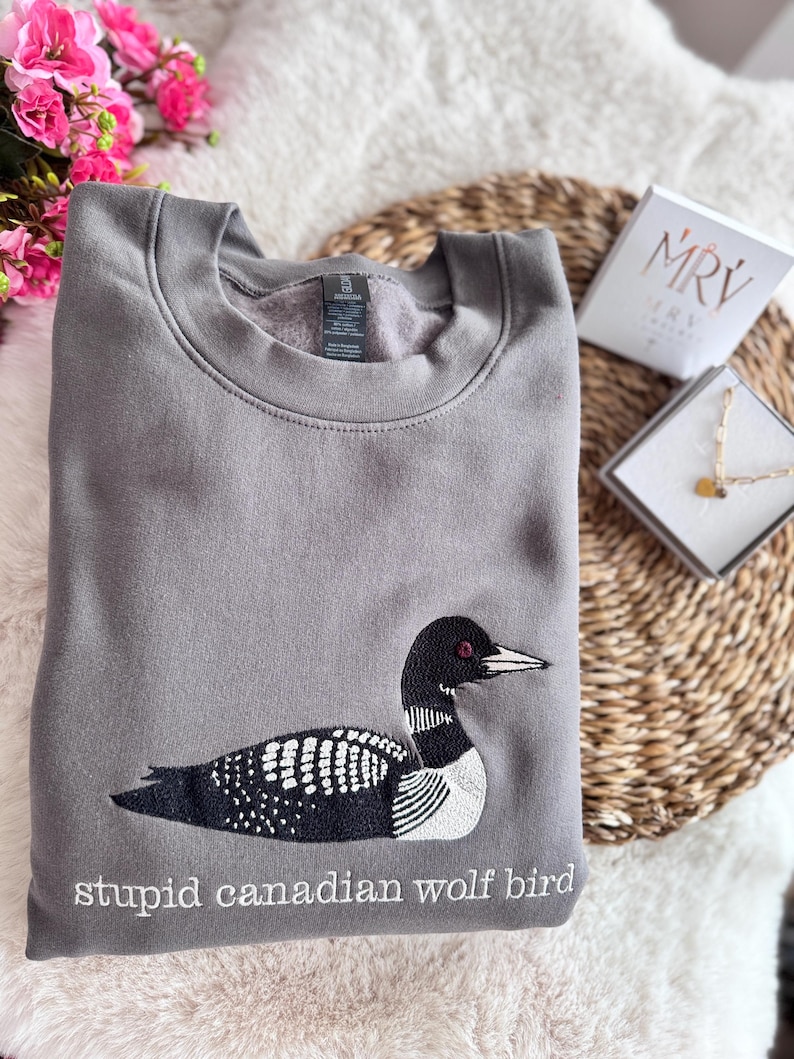 Pode incluir: Uma camisola cinzenta de gola redonda com um bordado de um mergulh&atilde;o em preto e branco. Por baixo da ave, o texto "stupid canadian wolf bird" est&aacute; bordado a branco. A camisola tem um interior em polar e uma gola padr&atilde;o.