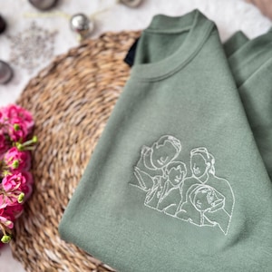 May include: An olive green crewneck sweatshirt featuring a white thread embroidered line art illustration of a family of four. The design depicts the outlines of two adults and two children positioned closely together in a portrait style.