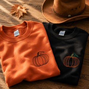 Puede incluir: Una sudadera naranja con una calabaza negra bordada y una sudadera negra con una calabaza naranja bordada, ambas con etiquetas Gildan. Un sombrero de vaquero marrón y una hoja de arce seca están colocados cerca sobre una superficie de madera.