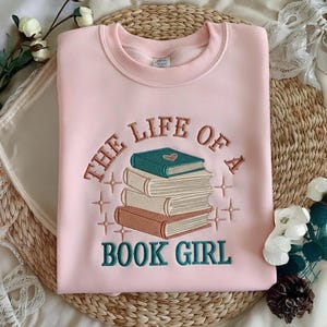 Könnte beinhalten: Ein hellrosa Sweatshirt mit Rundhalsausschnitt und einer Stickerei, die einen Stapel aus drei Büchern in den Farben Petrol, Creme und Braun zeigt. Der Text "THE LIFE OF A BOOK GIRL" ist bogenförmig um die Bücher gestickt und mit kleinen Sternen verziert.