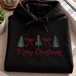 May include: A black hooded sweatshirt featuring an embroidered design of three green pine trees and two red bows. Below the trees, the text "Merry Christmas" is embroidered in red script. The hoodie includes a matching black drawstring at the neck.
