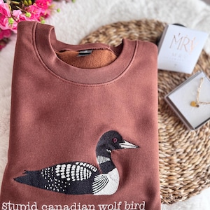 Pode incluir: Uma camisola castanha de gola redonda com um bordado de um mergulh&atilde;o em preto e branco. Por baixo da ave, o texto "stupid canadian wolf bird" est&aacute; bordado com fio branco. A camisola tem um interior em tecido polar macio.