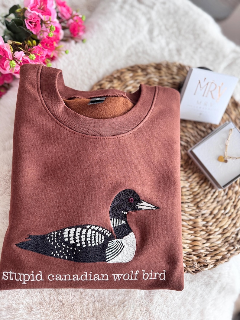 Pode incluir: Uma camisola castanha de gola redonda com um bordado de um mergulh&atilde;o em preto e branco. Por baixo da ave, o texto "stupid canadian wolf bird" est&aacute; bordado com fio branco. A camisola tem um interior em tecido polar macio.