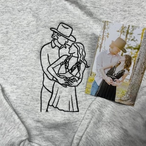 May include: A light gray heathered sweatshirt featuring a black embroidered line art illustration of two people embracing, one wearing a cowboy hat. Beside the embroidery is a rectangular photograph showing the same couple in an outdoor setting.