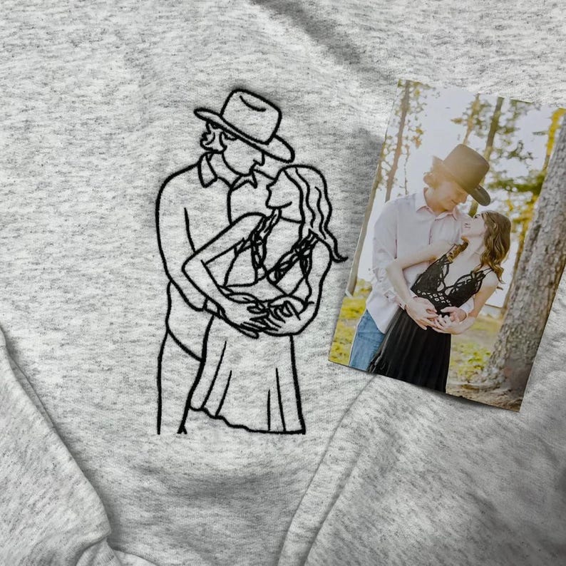 May include: A light gray heathered sweatshirt featuring a black embroidered line art illustration of two people embracing, one wearing a cowboy hat. Beside the embroidery is a rectangular photograph showing the same couple in an outdoor setting.