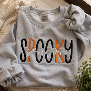 Puede incluir: Una sudadera gris claro de cuello redondo con el texto bordado SPOOKY en letras alternas negras y naranjas, con la palabra season escrita en cursiva blanca superpuesta sobre el centro del texto. La prenda tiene una etiqueta de la marca Gildan.