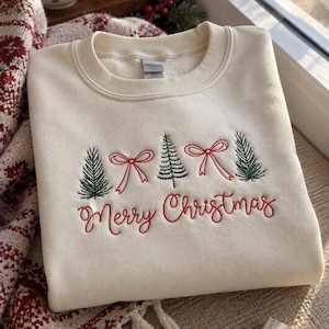 Könnte beinhalten: Ein cremefarbenes Sweatshirt mit Rundhalsausschnitt, das auf der Brust mit grünen Tannenbäumen und roten Schleifen bestickt ist. Unter der Stickerei ist der Schriftzug Merry Christmas in roter Schreibschrift eingestickt. Das Kleidungsstück hat gerippte Bündchen.