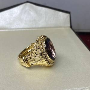May include: A gold ring with an oval, purplish-brown gemstone. The ring has an ornate, textured design with intricate details. The ring is set in a box with a neutral-colored interior.