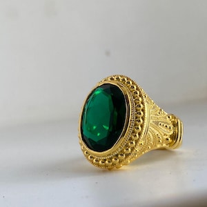 May include: A gold-colored ring with an oval, emerald-green gemstone. The ring has an ornate, textured design around the gemstone and along the band. The ring is set against a white background.