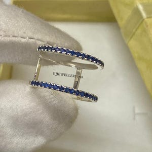 Puede incluir: Un anillo de doble banda de plata con dos filas paralelas de pequeñas piedras redondas de color azul intenso. El anillo tiene un sello 925 y el texto GJJEWELLERY1718 impreso en la banda interior. El diseño consta de dos bandas metálicas finas unidas por barras verticales.