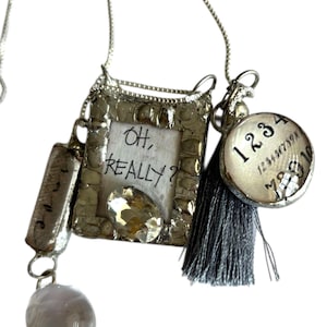 Può includere: Collana color argento con diversi ciondoli. I ciondoli includono un pezzo rettangolare, una cornice con la scritta "OH, REALLY?", un ciondolo rotondo con numeri, un nappa grigia e un globo grigio e bianco.