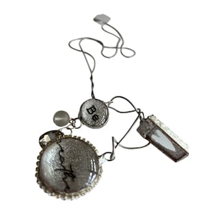 Può includere: Collana color argento con più ciondoli. La collana presenta un ciondolo rotondo con la parola "faith" in corsivo, un piccolo ciondolo circolare con la parola "Be" e un ciondolo rettangolare. Una piccola perla e un cristallo accentuano il design.