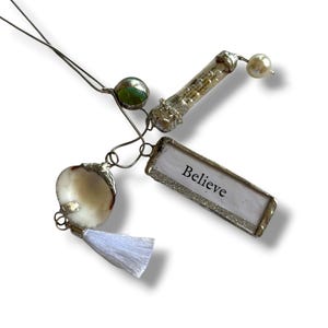 Può includere: Una collezione di ciondoli e pendenti fatti a mano. Include una conchiglia, un tubo di vetro, una perla verde e un pendente rettangolare con la scritta "Believe" in lettere argentate. Ogni ciondolo è collegato con filo d'argento e un nappa bianca.