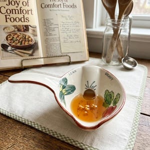 Puede incluir: Una salsera de cerámica con asa, llena de líquido y especias, se encuentra sobre un mantel individual a cuadros blancos y verdes. El recipiente está decorado con verduras pintadas y las palabras "LEAN" y "GRAVY". Un libro de cocina abierto titulado "The Joy of Comfort Foods" está en el fondo.
