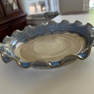 May include: A handmade ceramic pie dish with a wavy, fluted rim. The dish features a light brown center with concentric swirl patterns and a blue-glazed rim. The rim's edge has a darker blue accent. This dish is perfect for baking and serving pies.
