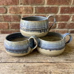 May include: Three ceramic mugs with a wide, low-profile shape and a single handle. The mugs feature a reactive glaze with a blend of blue, grey, and cream tones, showing a distinct drip pattern where the blue glaze overlaps the cream base.