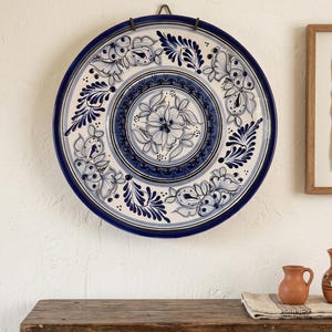 May include: A large, round decorative plate with a blue and white floral design. The plate is mounted on a wall and features a dark blue rim and intricate floral patterns in the center. The plate is approximately 12 inches in diameter.