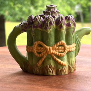 May include: A ceramic teapot shaped like a bundle of asparagus. The teapot is mainly green with purple asparagus tips and a tan rope design around the middle. The handle and spout are also green.