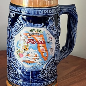 May include: A ceramic beer stein with a blue and orange color scheme. The stein features a map of Florida with illustrations of local landmarks. The handle has a tree branch design. The stein has a decorative rim and base.